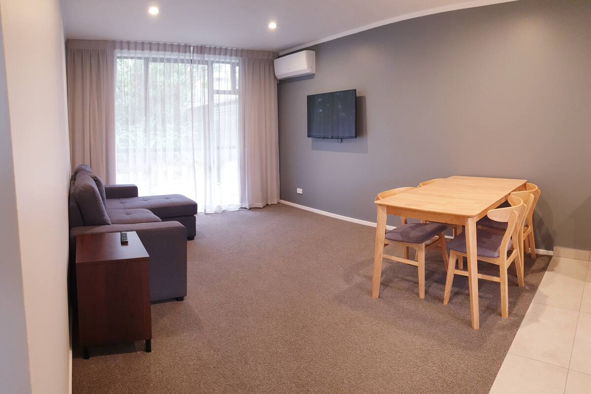 Uma sala de estar com sofá, televisão fixada na parede, mesa de jantar de madeira com cadeiras e piso coberto de carpete.