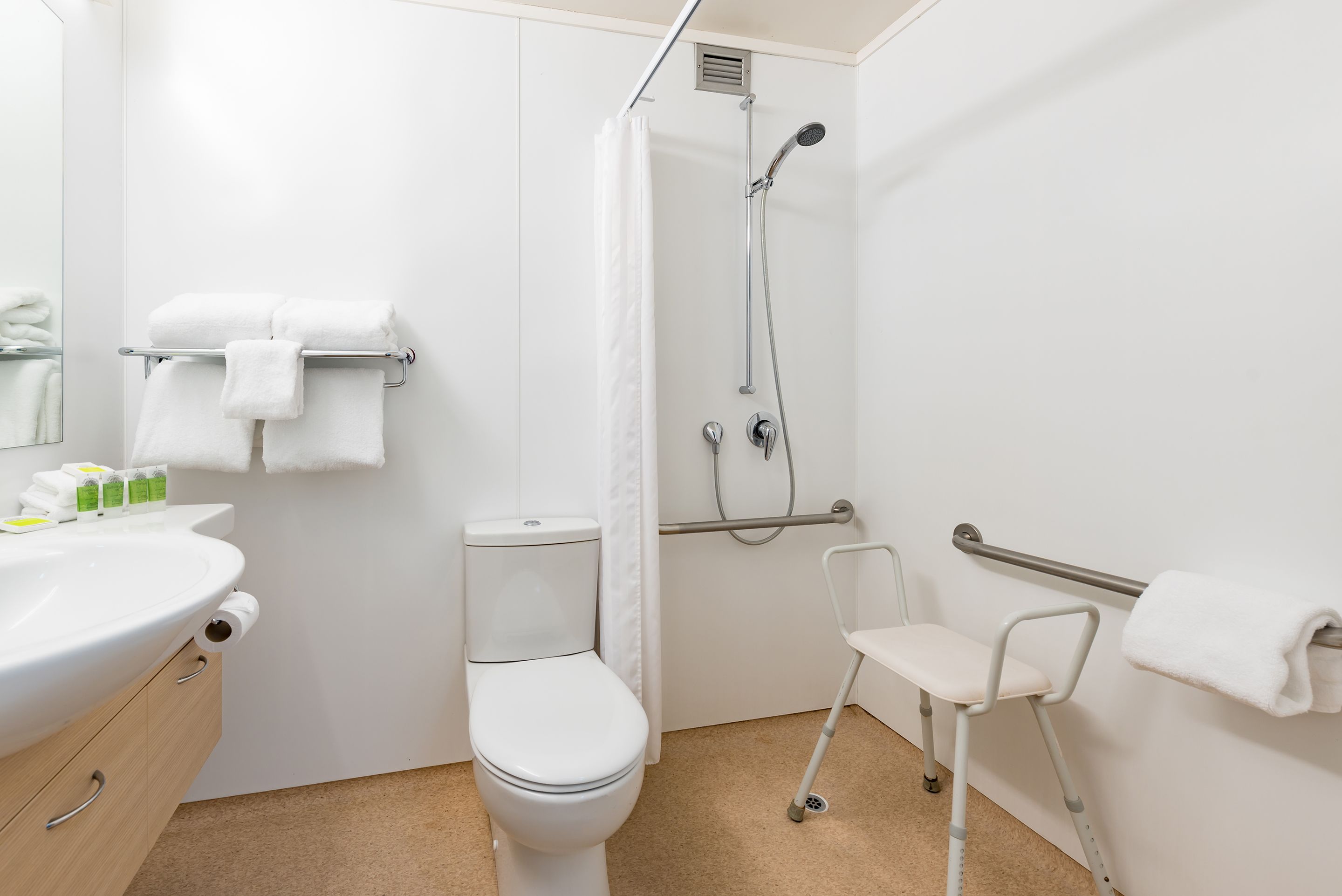 A bathroom with a toilet, shower, grab bar, and a chair.