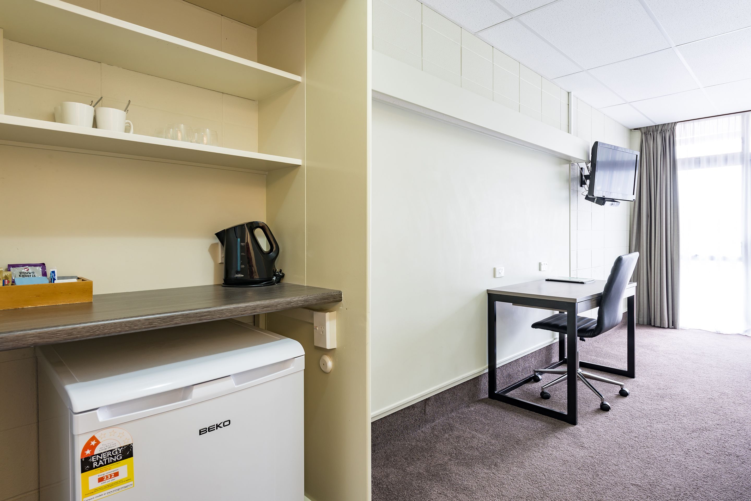 A hotel room with a kitchenette, a desk with a chair, a television mounted on the wall, and a window with curtains.