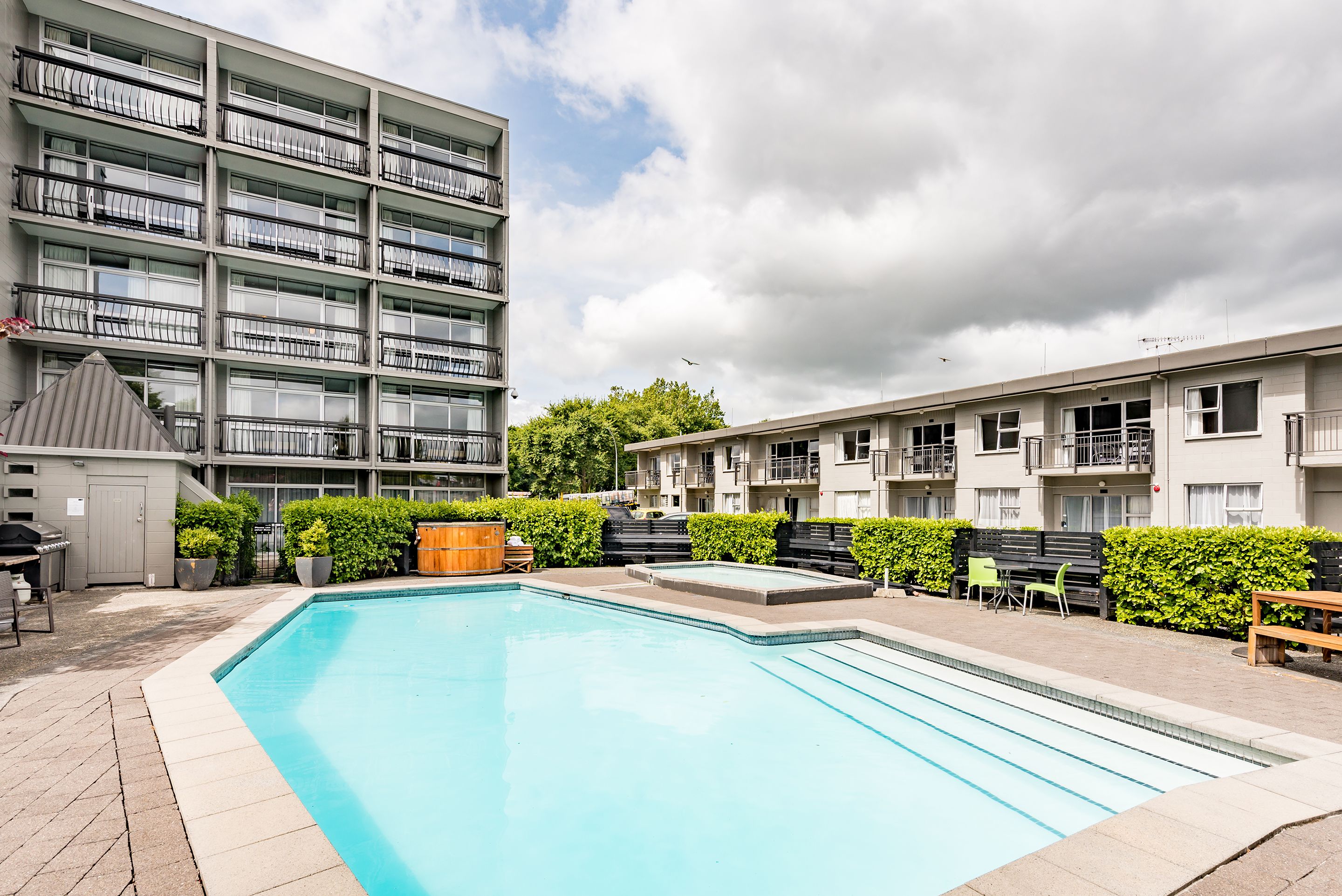 A swimming pool with a tiled deck, surrounded by a fence and greenery.