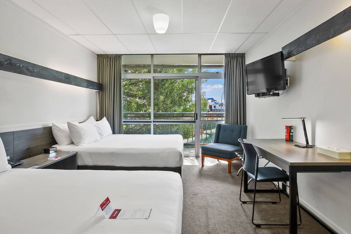 A hotel room with two beds, a desk, a chair, a TV, and a large window with a view of trees and buildings.