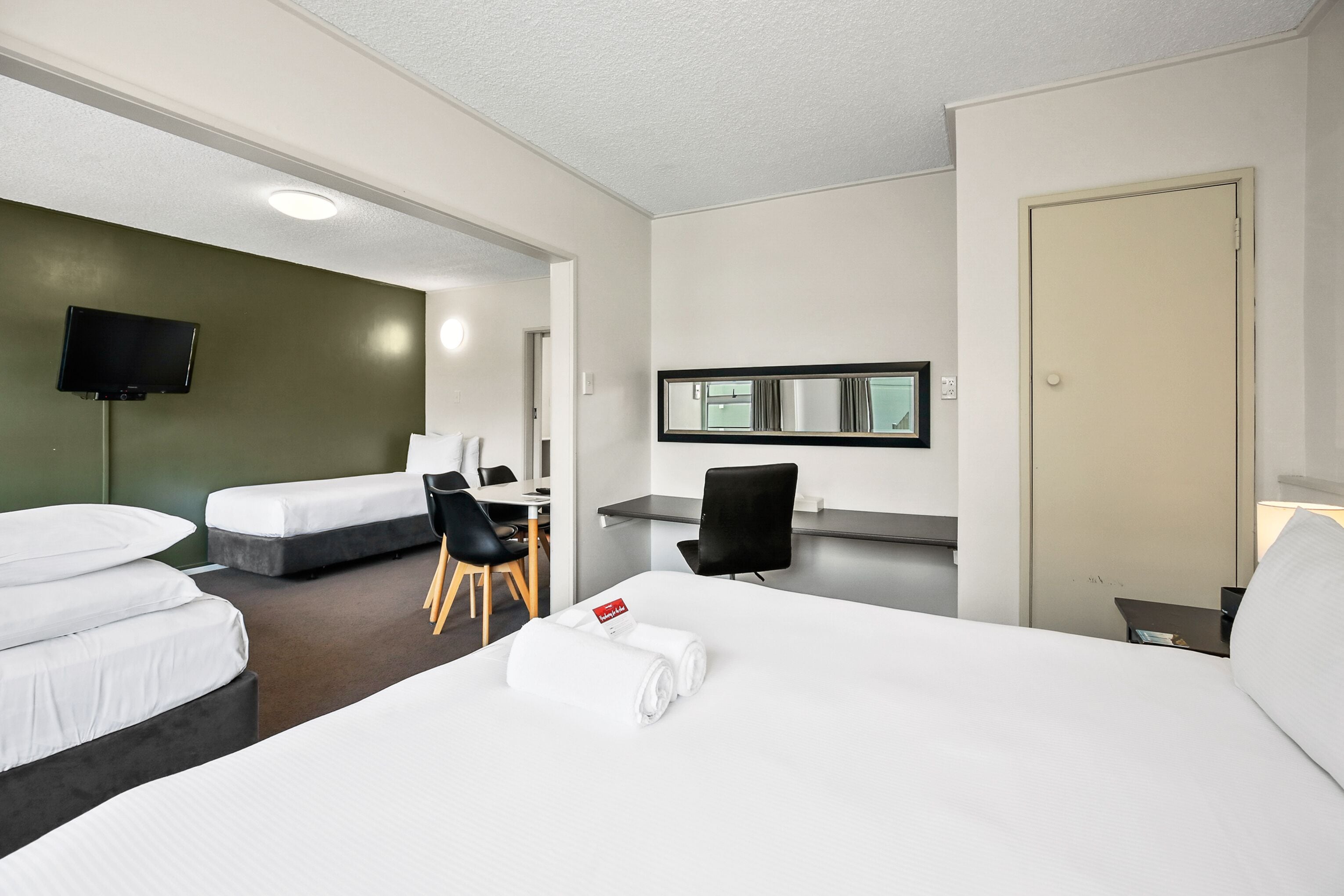 A hotel room with a large bed, two smaller beds, a desk with a chair, a TV, a mirror, and a door.