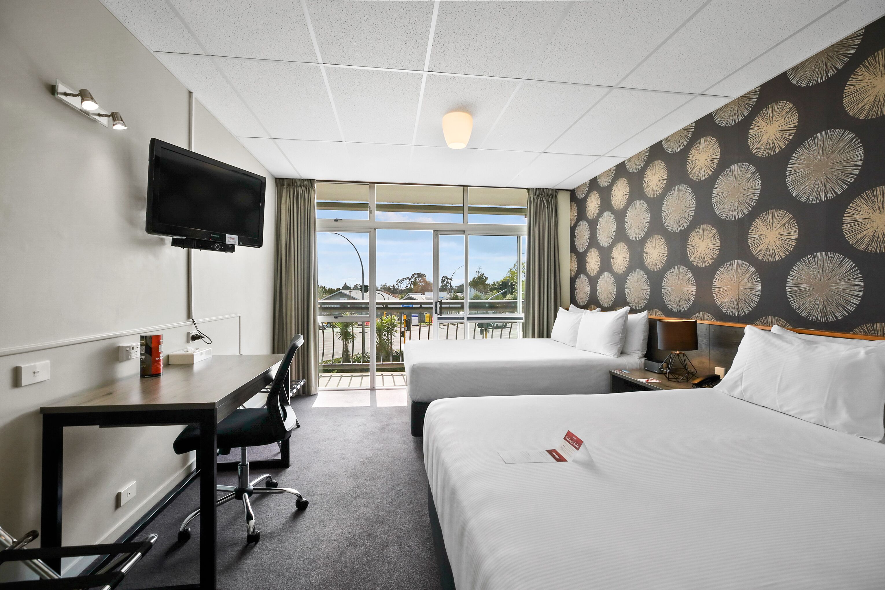 A hotel room with a desk, two beds, a TV, and a view of the outdoors.