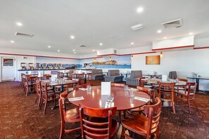 A spacious dining area with multiple round tables and chairs, a kitchen in the background, and a mural of a coastal scene.