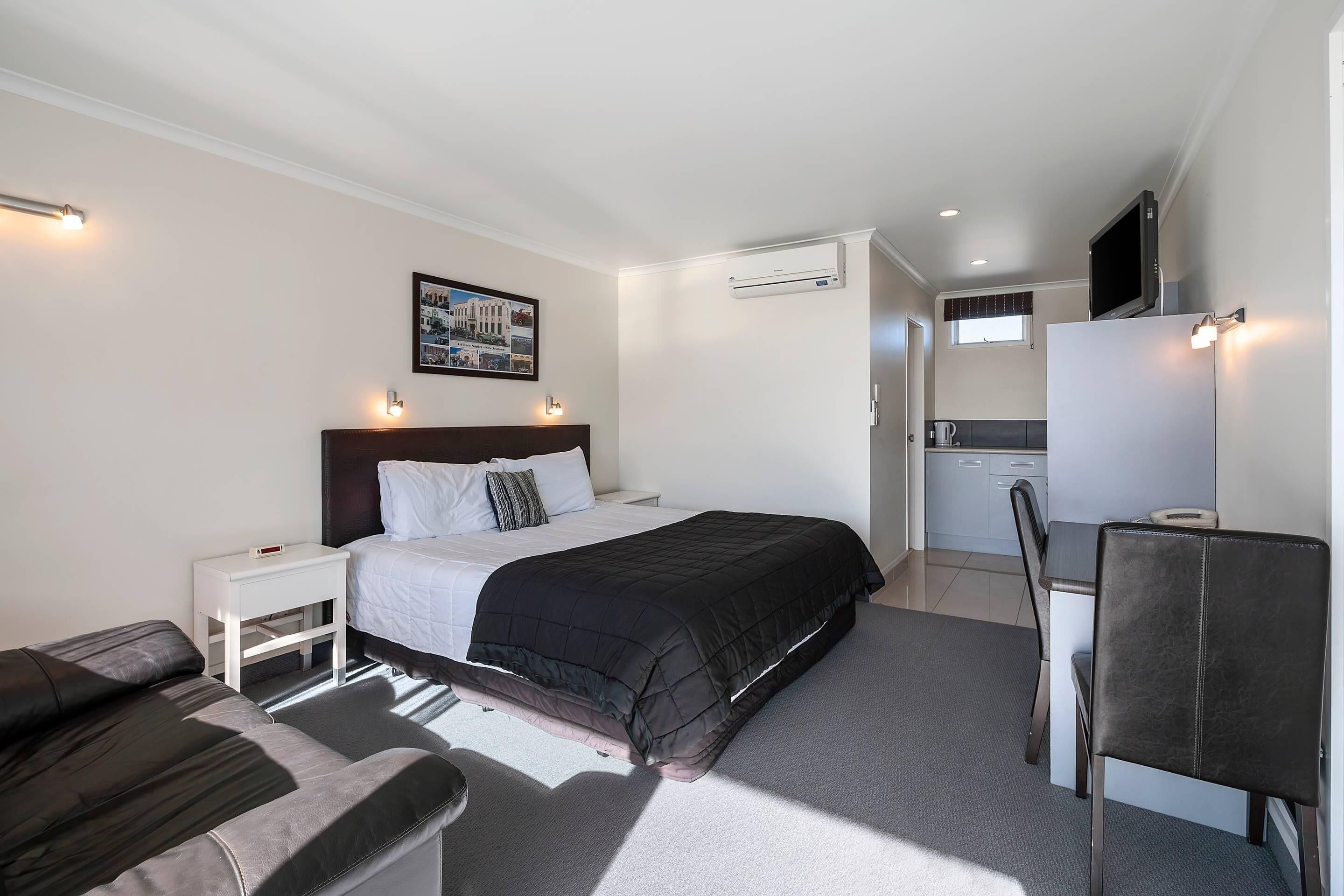 A bedroom with a bed, bedside table, and a desk with a chair. There is a kitchenette in the background.