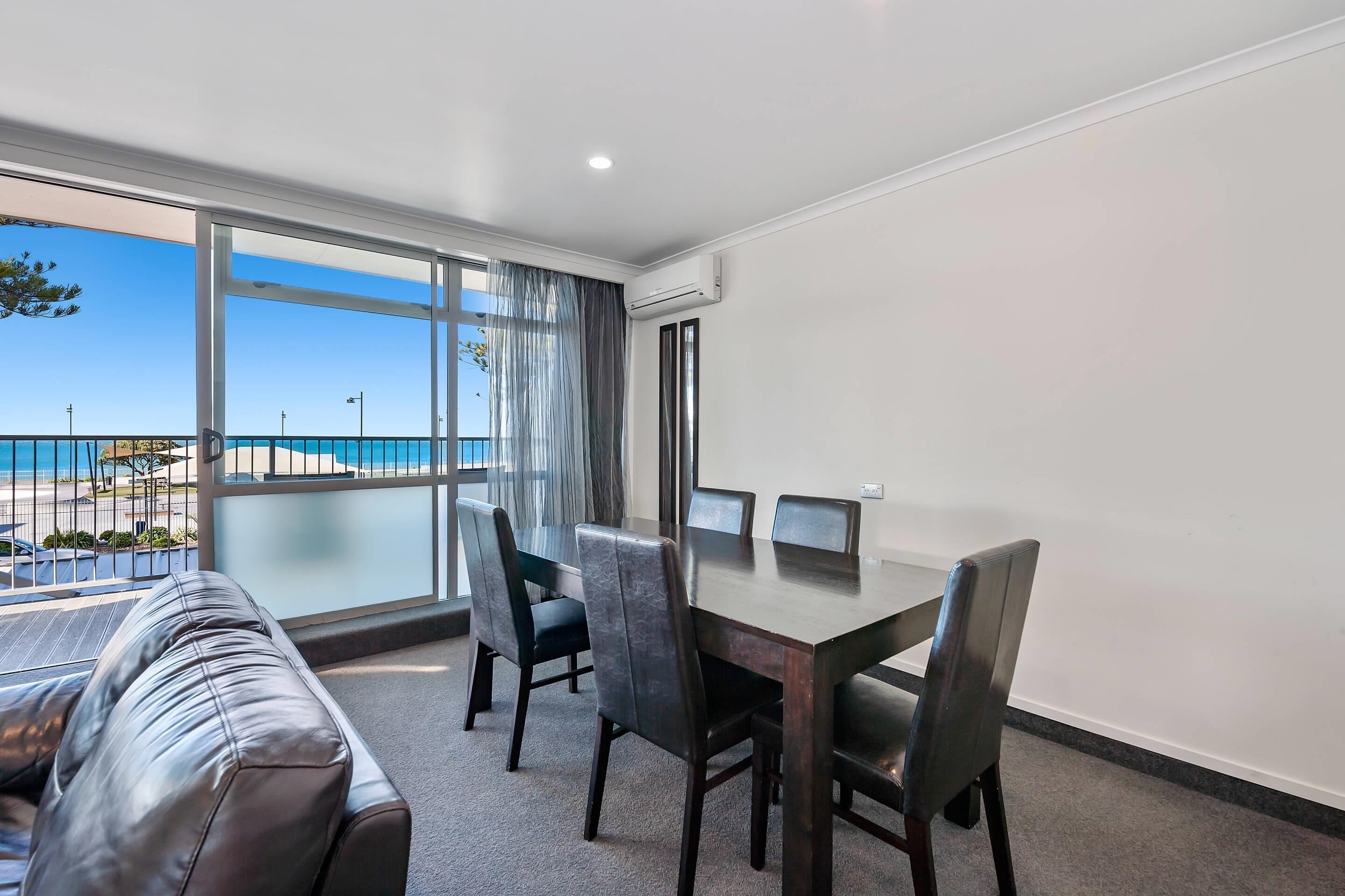 Ett modernt hotellrum med ett stort fönster som ger utsikt över stranden, ett matbord med stolar och en soffa klädd i läder.