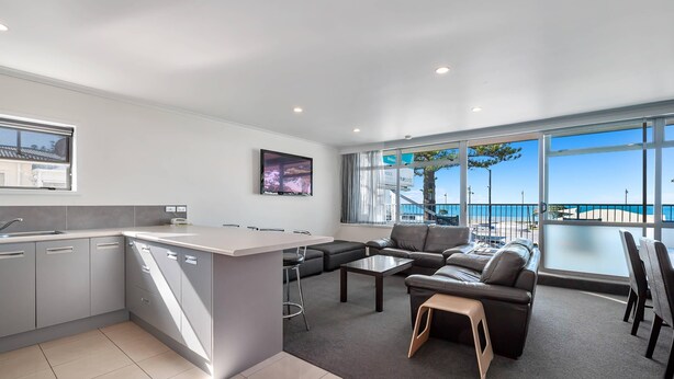 A modern kitchen with a central island, a dining area with a sofa and chairs, and large windows offering a view of the sea.