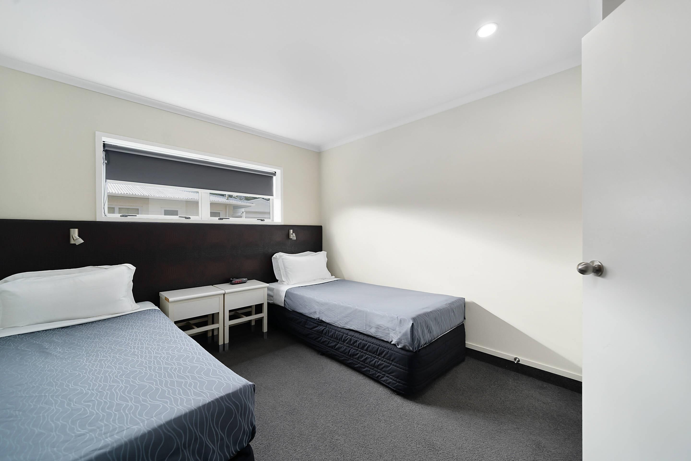 A small, clean room with two single beds, a small desk, and a window with blinds.
