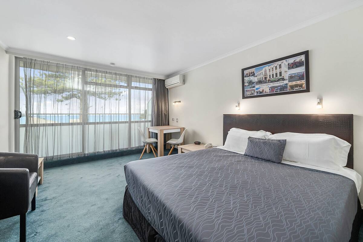 Habitación de hotel con una cama grande, un escritorio y una silla. Se ve una ventana con cortinas, un equipo de aire acondicionado y un cuadro en la pared.