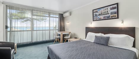Une chambre d’hôtel avec un grand lit, un bureau et une chaise. Il y a une fenêtre avec des rideaux, un climatiseur et un tableau encadré au mur.
