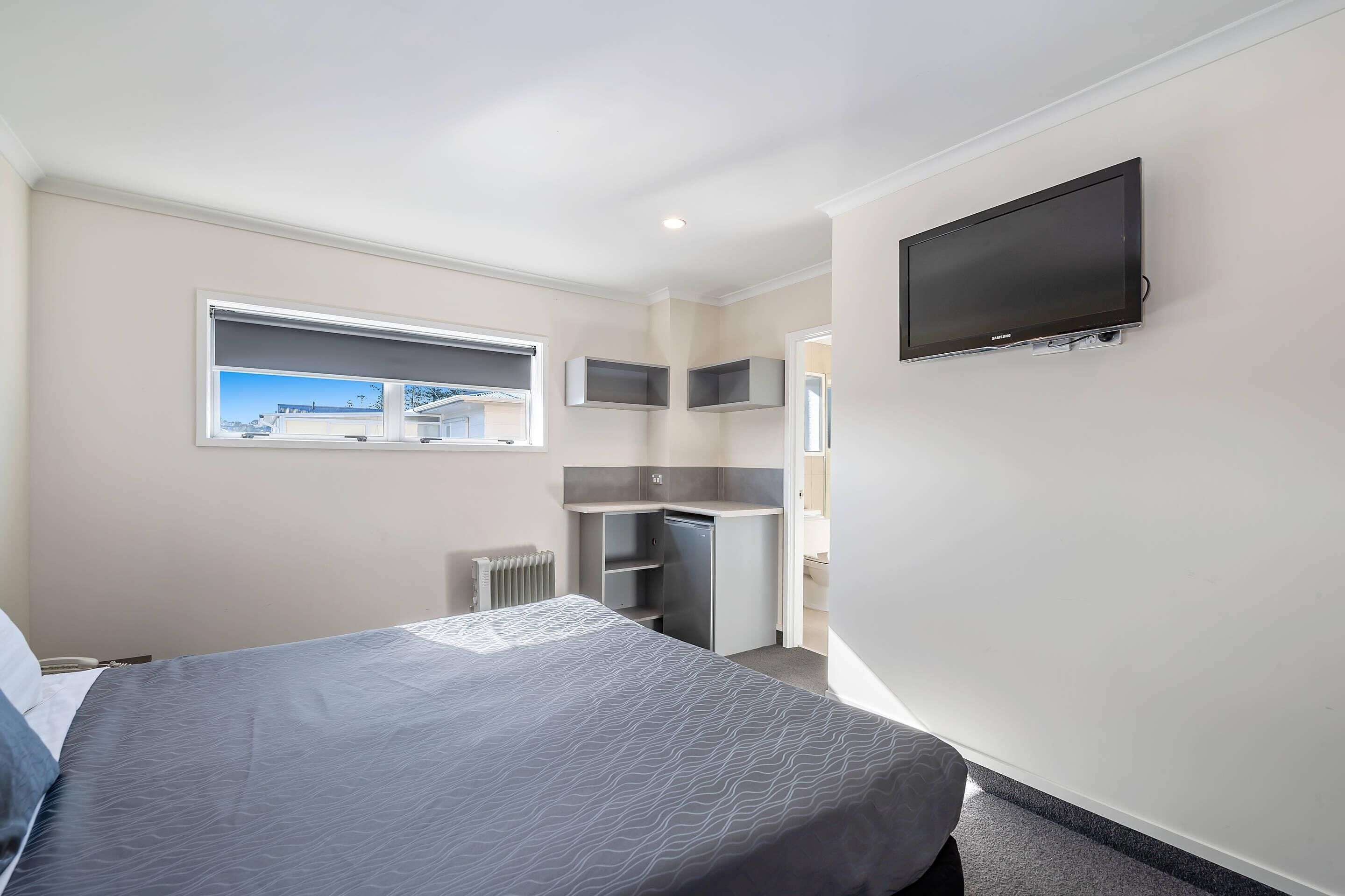 Een slaapkamer met een bed, een aan de muur bevestigde televisie, een raam en een ingebouwd bureau met planken.