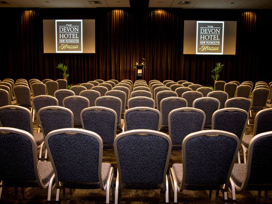 Ein Konferenzraum mit Reihen von Stühlen und zwei Bildschirmen, die die Marke 'The Devon Hotel New Plymouth Heritage' zeigen.