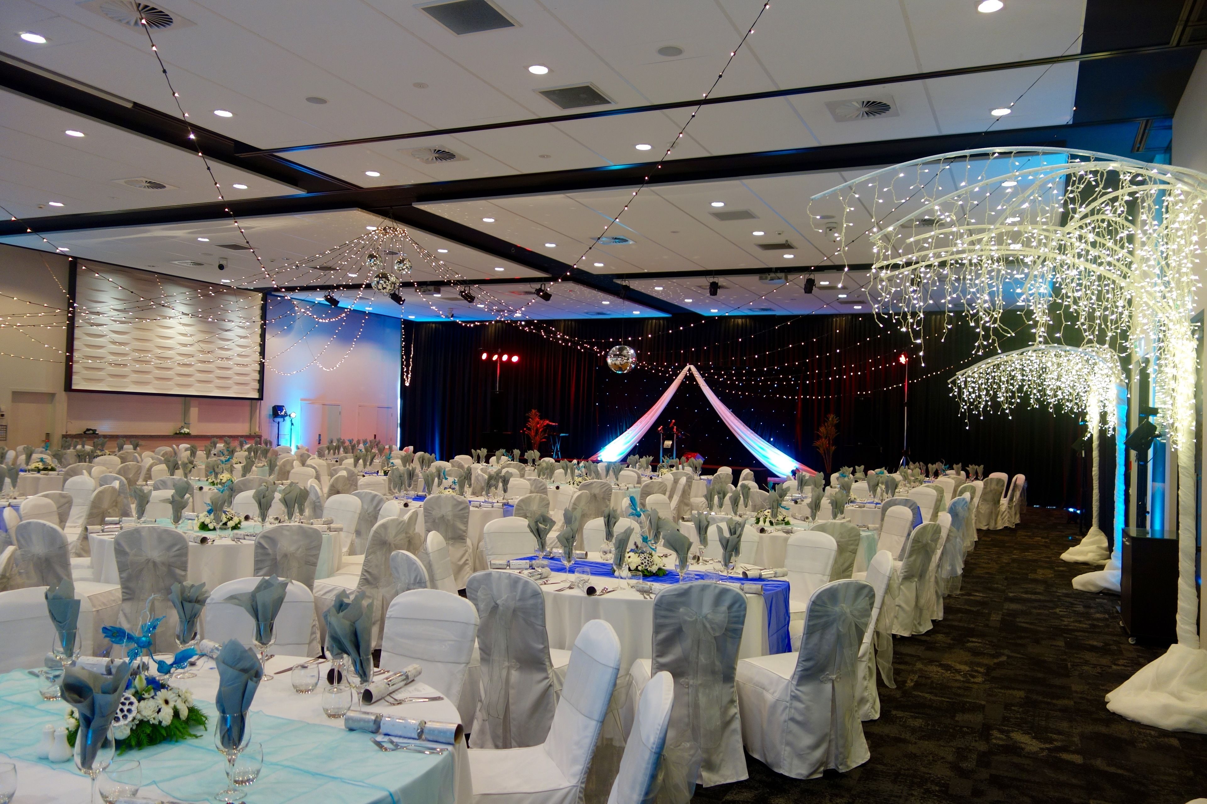 A banquet hall with rows of tables set for a formal event, featuring white tablecloths and chairs with white and blue sashes.