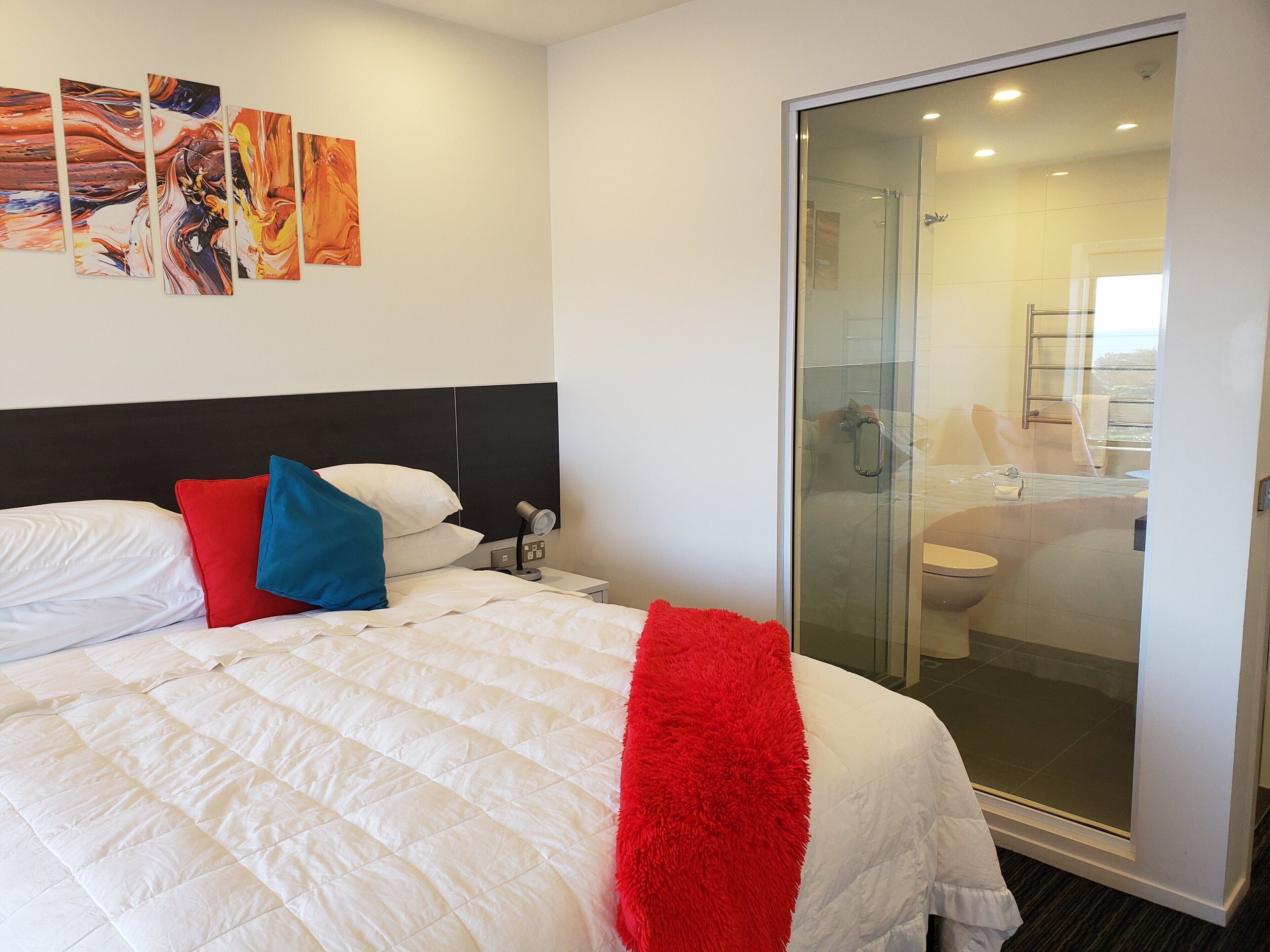 A hotel room with a bed, a nightstand, a bedside table with a lamp, a glass shower enclosure, and a bathroom visible through the glass door.