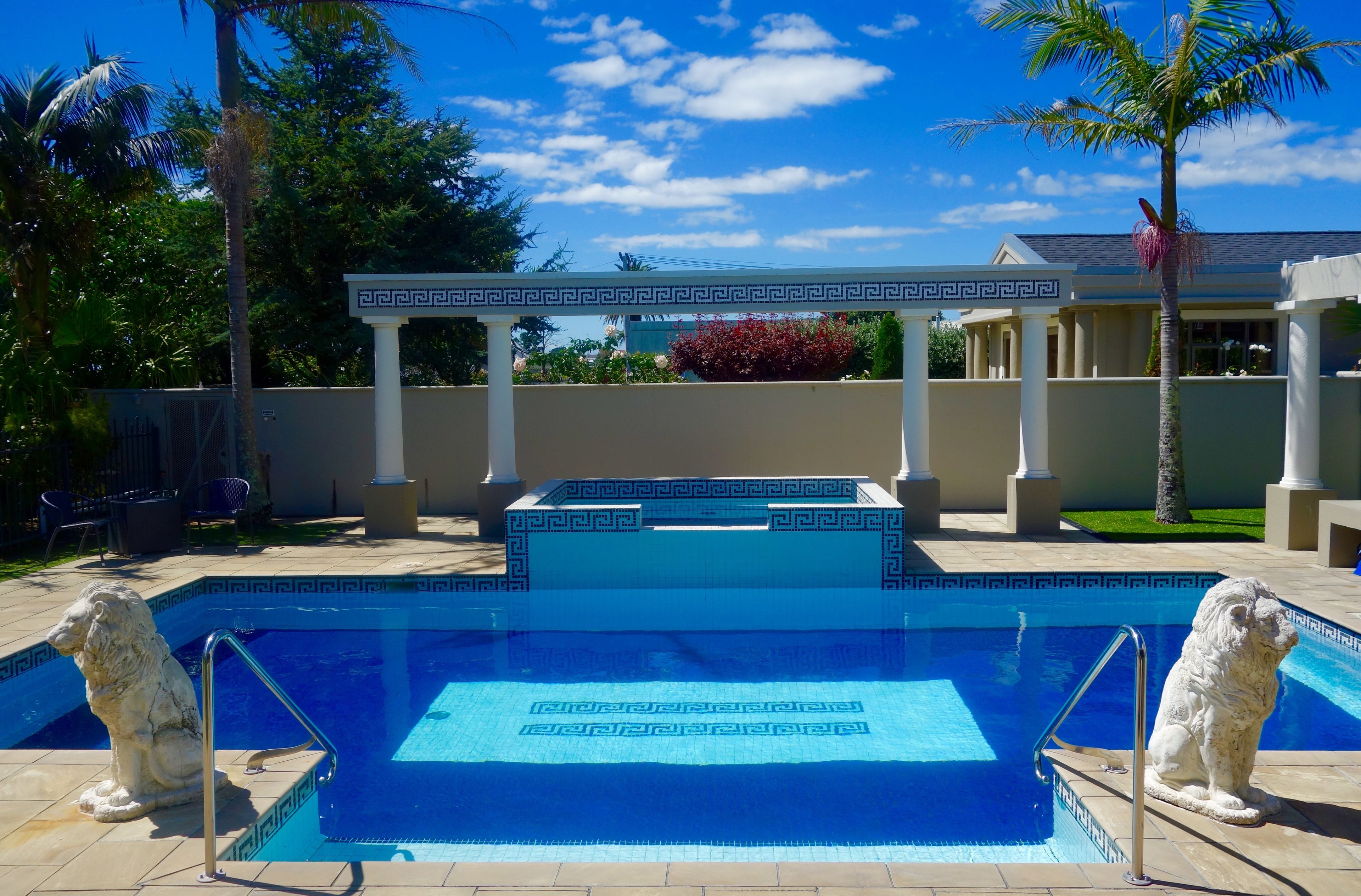 A swimming pool with a geometric tile pattern, two stone lion statues, a pergola with a Greek key design, and a well-maintained garden with palm trees and a red flowering bush.