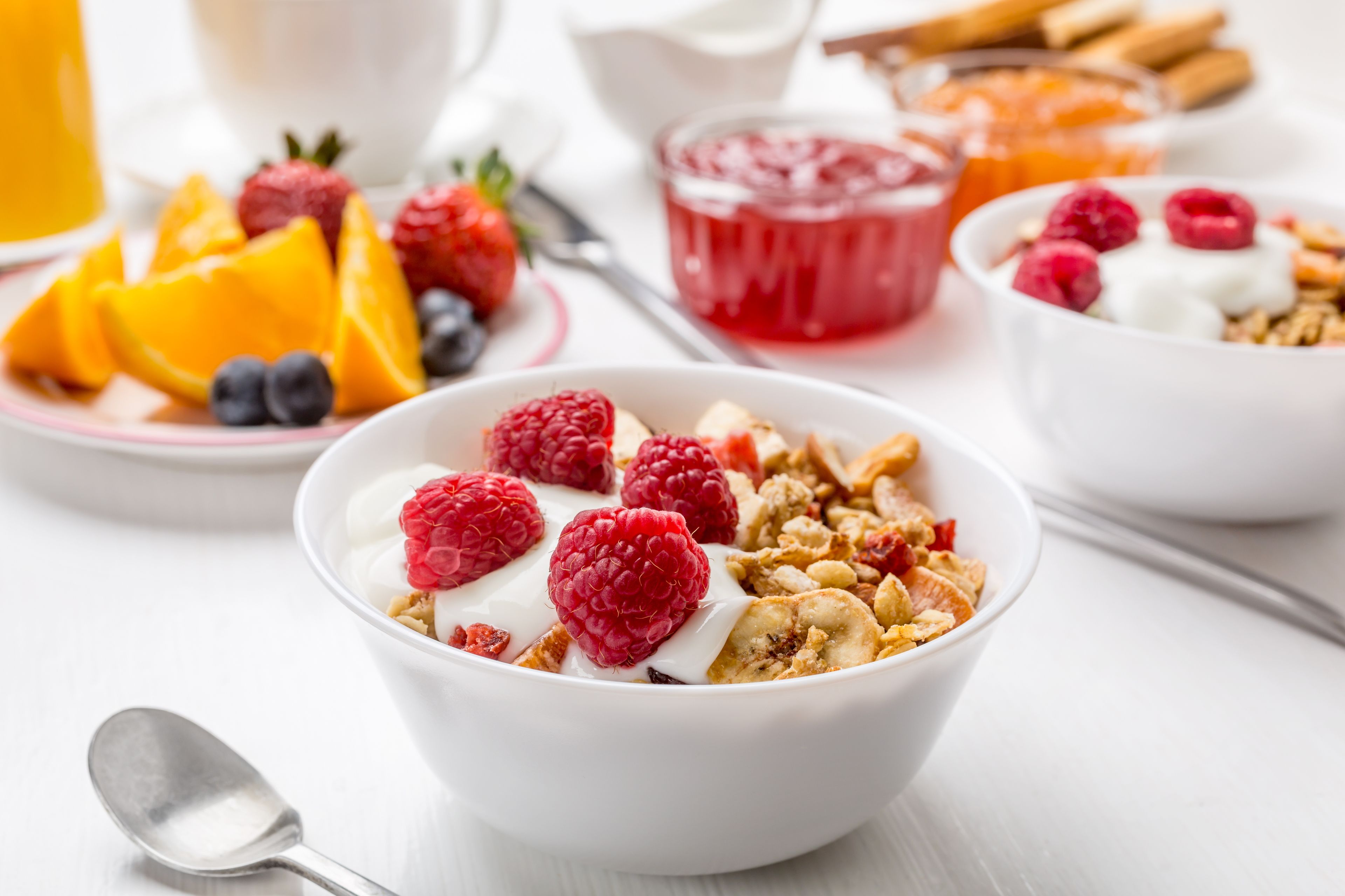 Een ontbijttafel met bakjes yoghurt en granola, belegd met frambozen en blauwe bessen, vergezeld van een glas sinaasappelsap en een schaal fruitsalade.