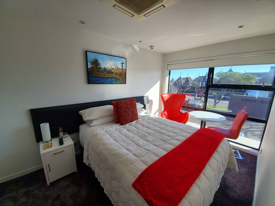 A hotel room with a bed, a nightstand, a red chair, a small table, and a large window offering a view of the outside.