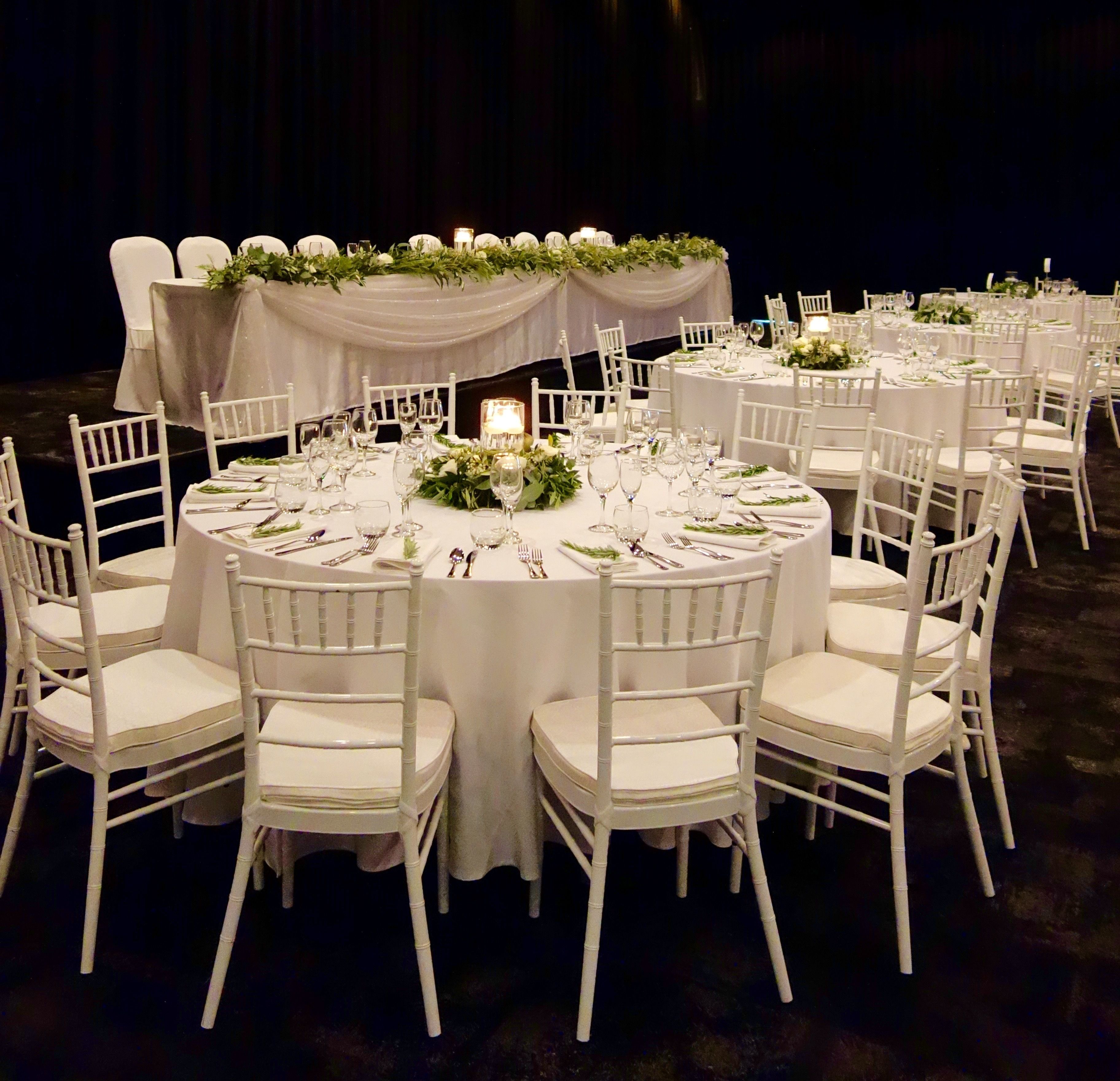 A banquet hall with round tables set for dining, white chairs, and floral centerpieces.