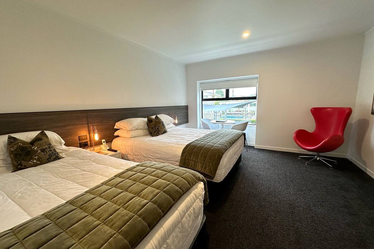 Habitación de hotel con dos camas, una silla roja y una ventana con vista a una piscina y un puente.