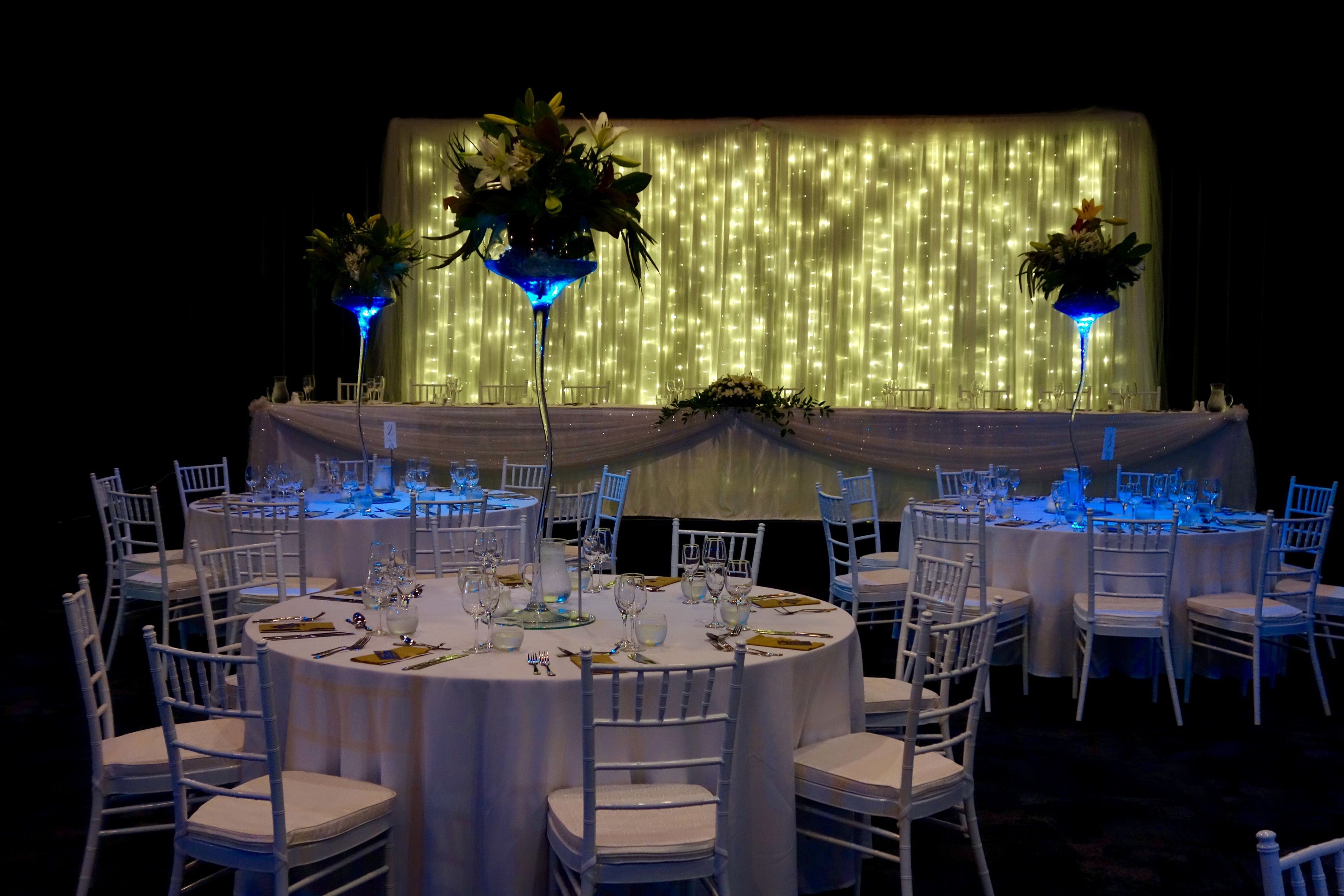 A banquet hall with round tables set for dining, white chairs, and blue ambient lighting.