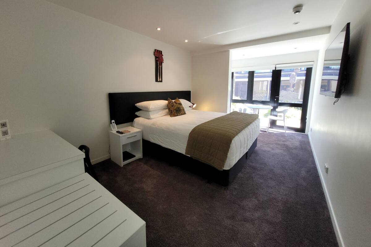 Una habitación de hotel moderna con una cama grande, una mesita de noche con teléfono, un escritorio pequeño y un ventanal con vistas al exterior.