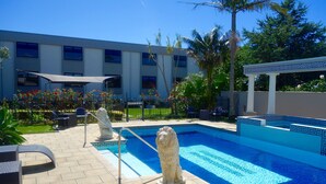 Una piscina con una estatua decorativa de león, mobiliario exterior y un edificio al fondo.