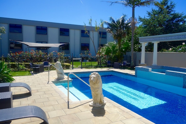 Una piscina con una estatua decorativa de león, mobiliario exterior y un edificio al fondo.