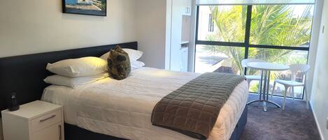 A neatly made bed with a quilted cover, a pillow, and a blanket. There is a small bedside table with a phone and a drawer. A painting hangs on the wall above the bed.