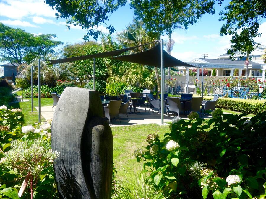 An outdoor patio area with a table and chairs under a shaded canopy, surrounded by lush greenery and flowering plants.