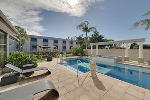 Zona de piscina con sillones de descanso, una pérgola y un edificio al fondo.