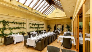 Un restaurante con mesas largas y sillas de mimbre, un techo alto con tragaluces y follaje decorativo en las paredes.