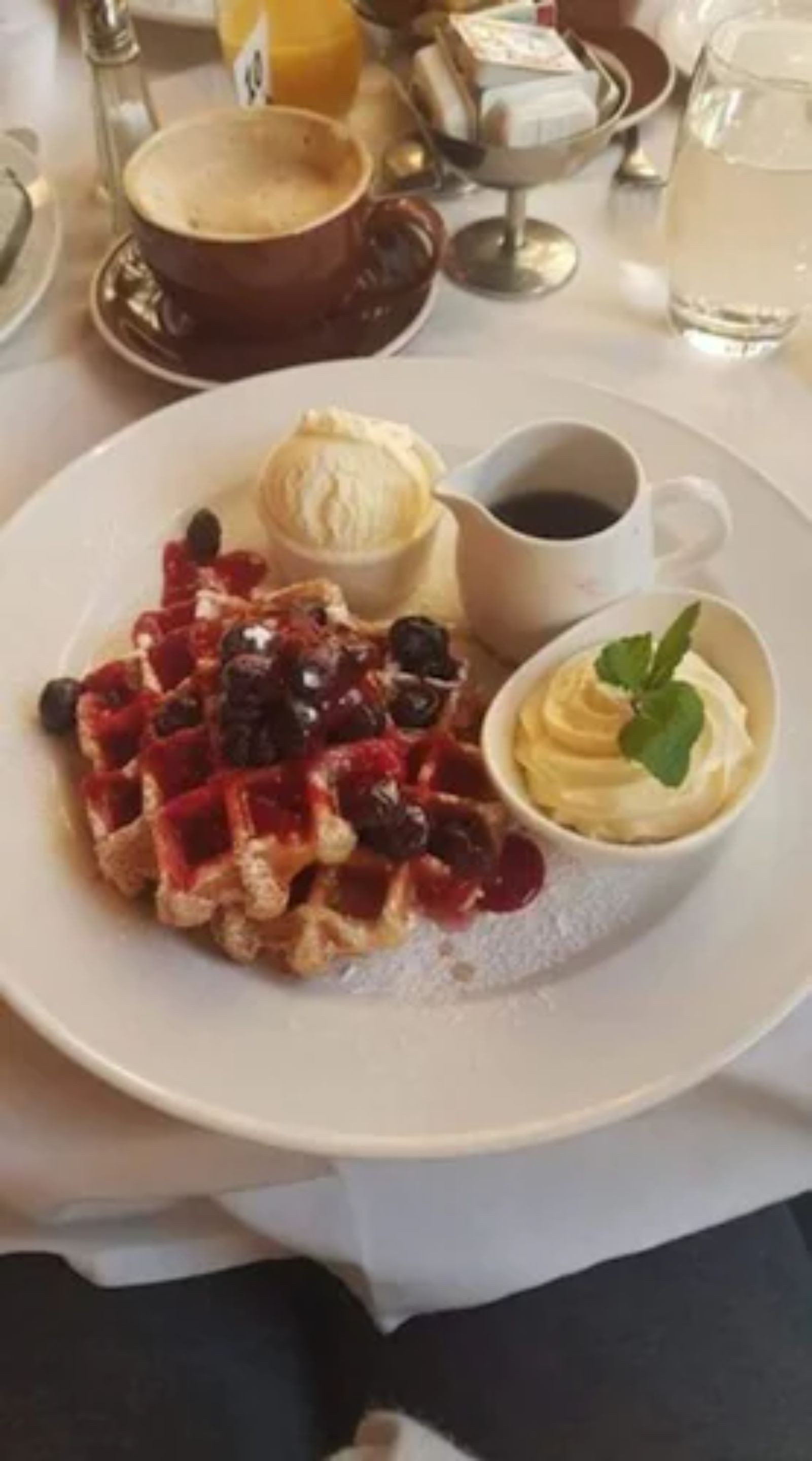 Un plato con waffles cubiertos de bayas y crema batida, una taza de café y un recipiente pequeño de crema batida con una hoja de menta.