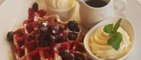 Un plato con waffles cubiertos de bayas y crema batida, una taza de café y un recipiente pequeño de crema batida con una hoja de menta.