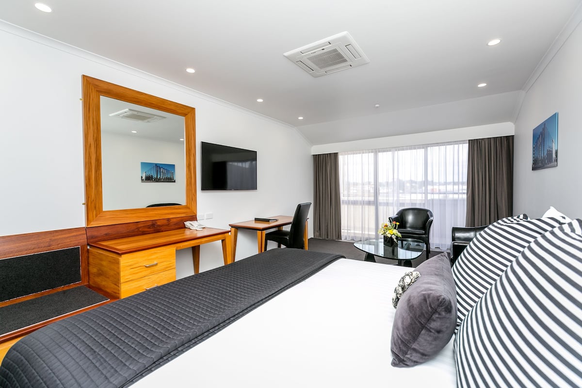 Ein Hotelzimmer mit einem Bett, einem Schreibtisch, einem Sessel, einem Fernseher und einem Vorhangfenster.