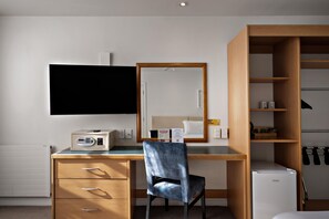 Habitación de hotel con un escritorio de madera, una silla, un televisor, un espejo, un armario y un refrigerador pequeño.