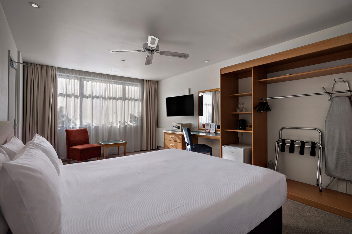 A hotel room with a bed, a desk, a chair, a television, a wardrobe, and a ceiling fan.