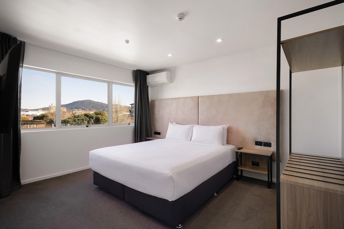 A hotel room with a large bed, a bedside table, a mirror, a window with a view of a mountain, and an air conditioning unit.