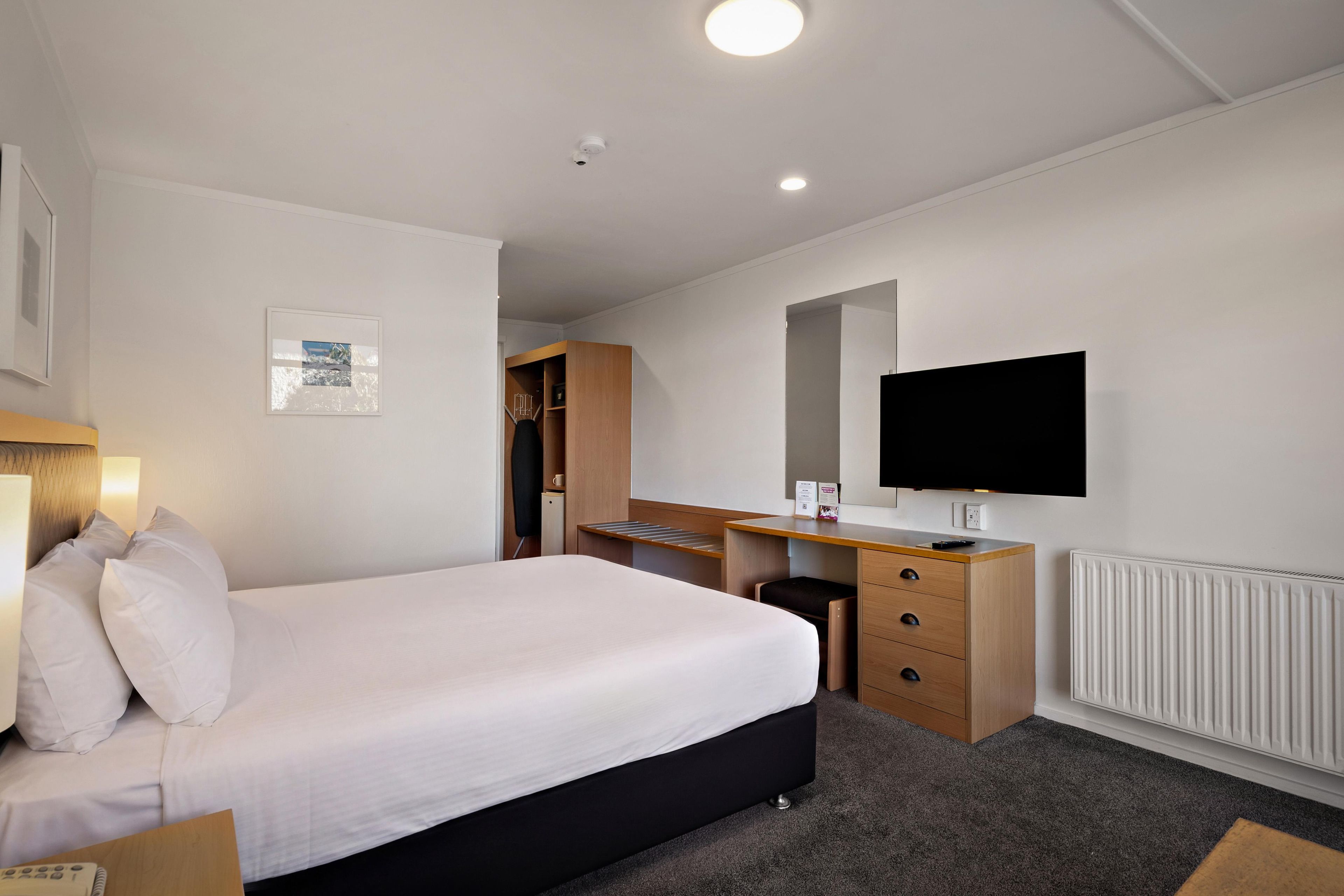 A hotel room with a bed, a desk, a chair, a television, and a radiator.