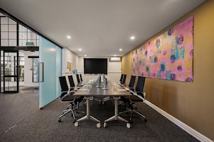 Uma sala de conferências com uma mesa comprida, cadeiras, uma tela de projetor e uma obra de arte abstrata colorida na parede.