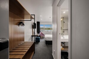 A hotel room with a wooden bench, a bed, a desk, a chair, and a view of the outdoors.