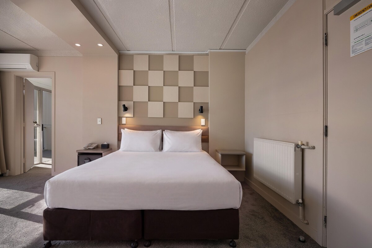 A hotel room with a large bed, bedside tables, a radiator, and a door with an emergency evacuation sign.