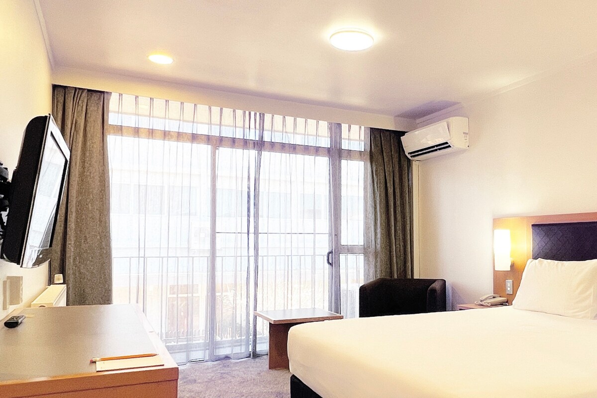 A hotel room with a bed, a desk, a chair, a small table, and a window with curtains.