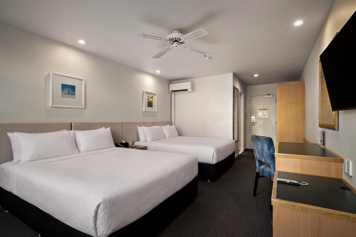 A hotel room with two beds, a desk, a chair, a ceiling fan, and an air conditioning unit.