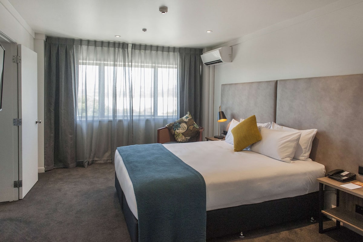 A hotel room with a large bed, a bedside table with a lamp, a window with curtains, and an air conditioning unit.