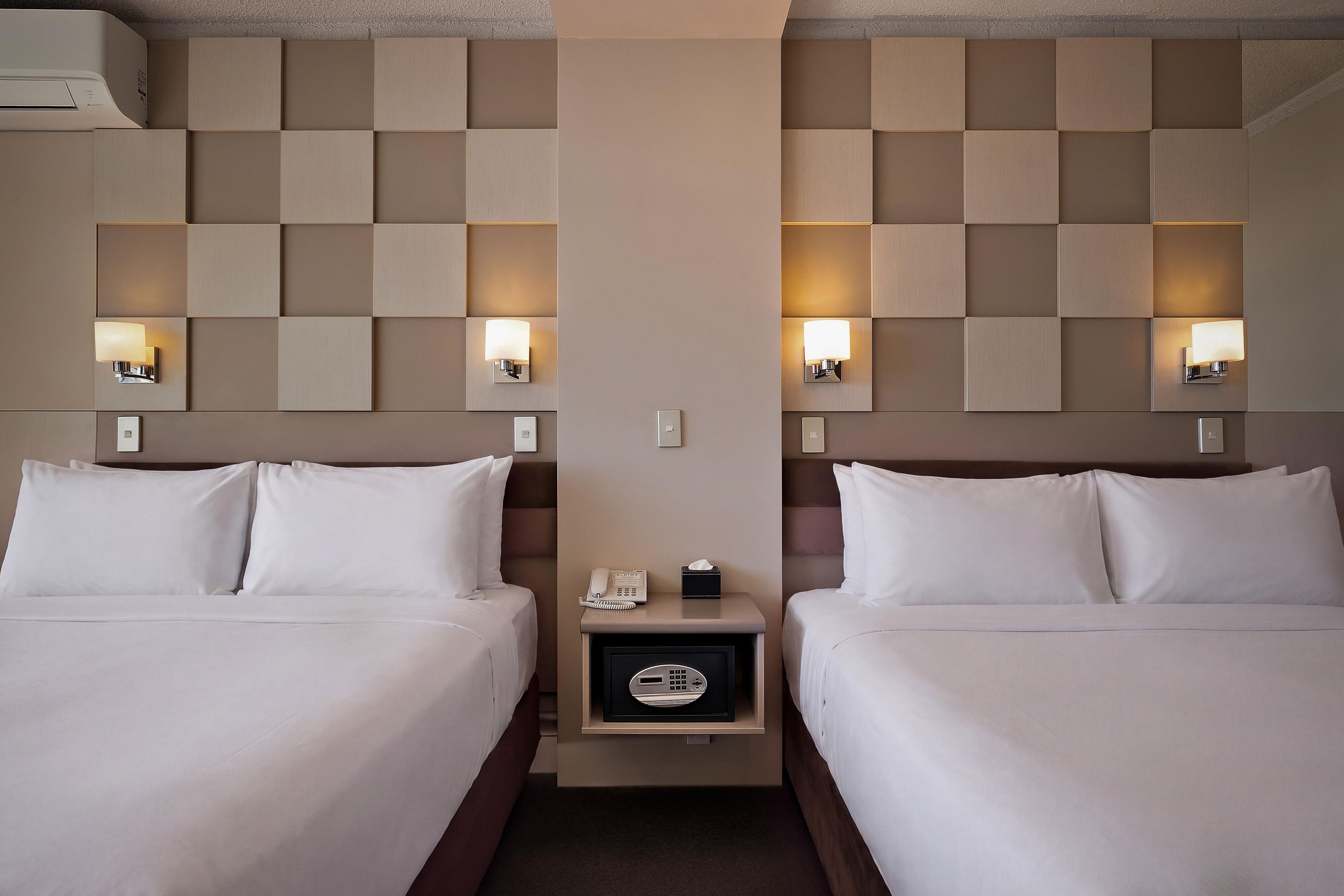 A hotel room with two beds, a nightstand with a phone and a tissue box, and wall-mounted lamps.