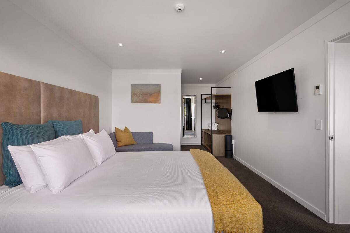 A modern hotel room with a large bed, a sofa, a TV, and a mirror.