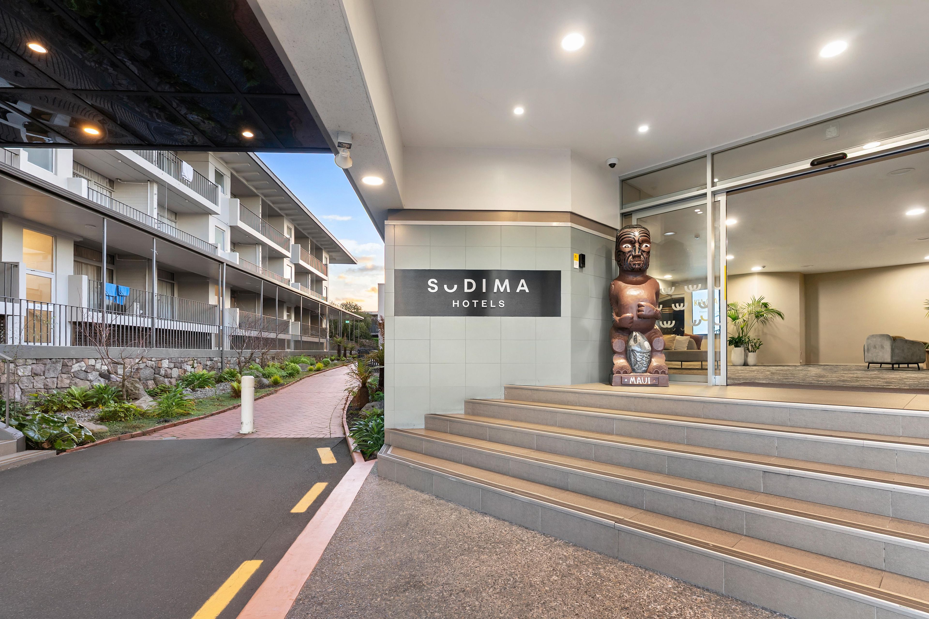 A hotel entrance with a sign for 'Sudima Hotels', a large statue, and a staircase leading to the lobby.