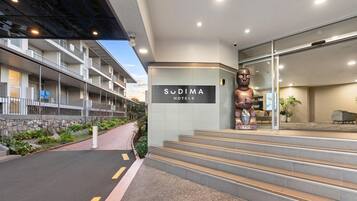 Een hotelentree met een bordje voor 'Sudima Hotels', een groot standbeeld en een trap naar de lobby.