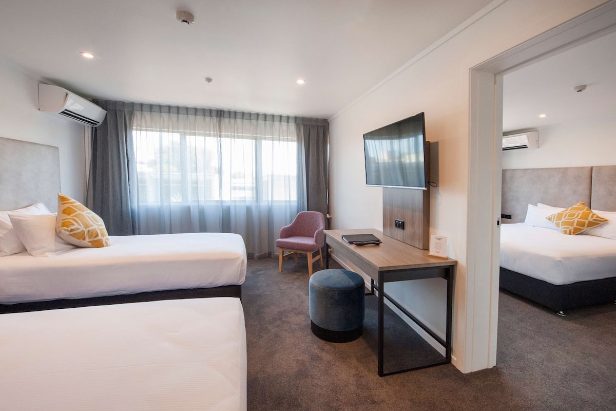 A hotel room with two beds, a desk, a chair, a TV, and an air conditioning unit.