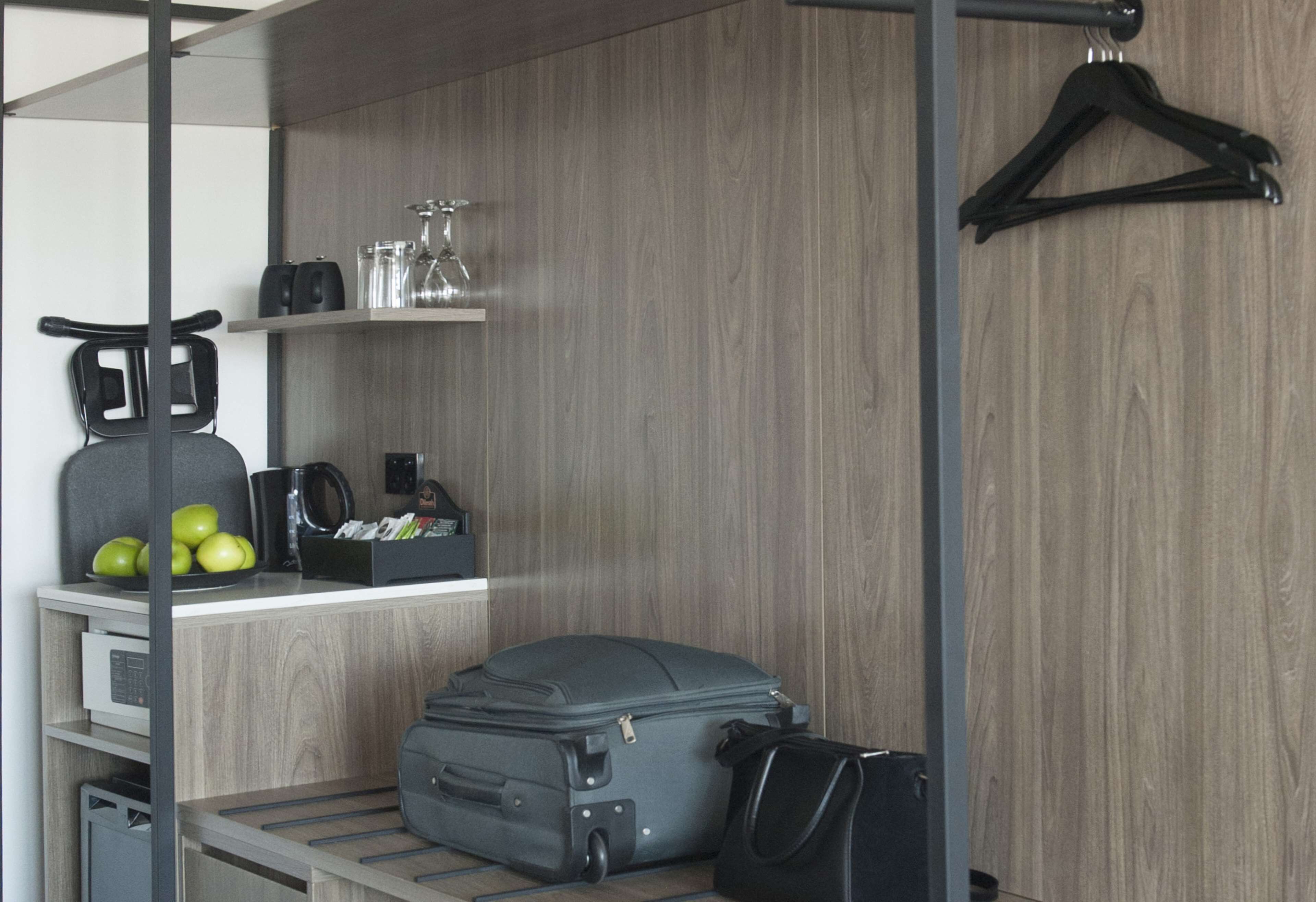 A compact hotel room with a wooden wall, a luggage rack, a small desk with a kettle, a coffee maker, and a few apples.